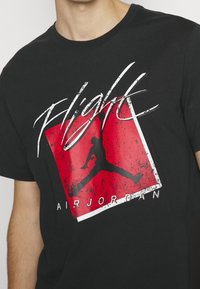 Černé krátké rukávy bavlněné tričko s červenou a bílou grafikou skákající postavy a textem "Flight AIR JORDAN" v tučném písmu.