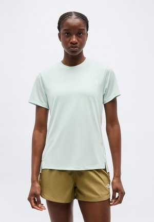 Γυναίκα με πλεγμένα μαλλιά που φοράει ανοιχτοπράσινο κοντομάνικο μπλουζάκι και κοντό χακί σορτς, στέκεται μπροστά σε μία απλή λευκή επιφάνεια.