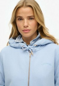 Sudadera con capucha y cremallera en azul claro, con cordón ajustable, herrajes dorados y logo bordado. Textura suave, combinada con una camisa con cuello debajo.