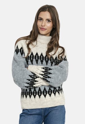 Kobieta o długich brązowych włosach, ubrana w szarą i kremową wzorzystą dzianinową bluzę oraz niebieskie jeansy, stojąca z założonymi ramionami.