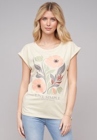 T-shirt couleur crème avec des manches courtes roulées, présentant un graphique floral en rose, vert et gris, et le texte "VIVEZ SIMPLE ET ÉPANOUISSEZ-VOUS SAUVAGEMENT."