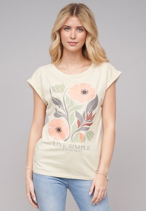 T-shirt couleur crème avec des manches courtes roulées, présentant un graphique floral en rose, vert et gris, et le texte "VIVEZ SIMPLE ET ÉPANOUISSEZ-VOUS SAUVAGEMENT."