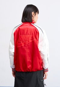 Raudona ir balta satino bomber tipo striukė su juodais akcentais. Pasižymi raukinėtais rankogaliais, aukštu apykakle ir reljefiniu "Nike" logotipu ant nugaros.