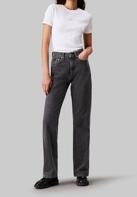 Bílý tričko s krátkým rukávem a logem, v kombinaci s černými džínami s vysokým pasem a rovnými nohavicemi, na černých botách, stojící na jednobarevném pozadí.