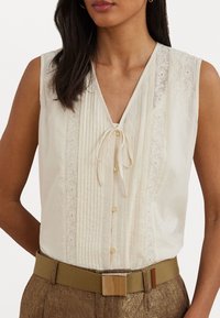 Ivory sleeveless blouse with a V-neck, pleated design, lace trim accents, and a front tie. Features small button closures and lightweight fabric.