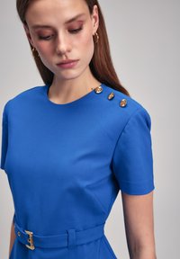 Blue dress with short sleeves, round neckline, fitted waist, gold button accents on shoulder, and a matching belt. Smooth, structured fabric.