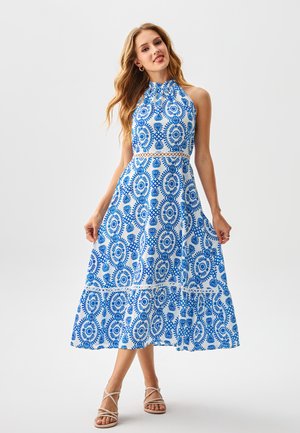 Vrouw draagt een mouwloze witte jurk met halternek en blauwe circulaire bloemmotieven, en beige sandalen met bandjes, staand op een witte achtergrond.