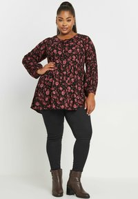 Tunica nera a fiori con maniche lunghe, caratterizzata da un orlo peplum arricciato, stampa di fiori rosa e dettagli con bottoni. Indossata con jeans neri e stivali marroni.