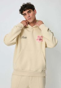 Sweat à capuche beige avec une poche avant, présentant une impression de fleur rose sur le côté gauche et le texte "Jardin de Provence" en noir. Texture lisse.
