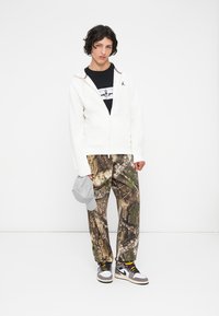Felpa bianca con zip, t-shirt nera con grafica, pantaloni mimetici con motivi marroni e verdi, cappellino grigio e sneakers marroni e bianche.