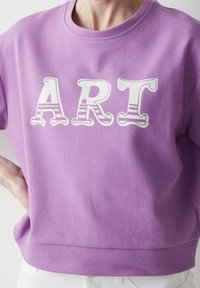 Lila bomullströja med vit text "ART" som har varierande mönster och former. Rund halsringning och ribbat fåll. Avslappnad passform.