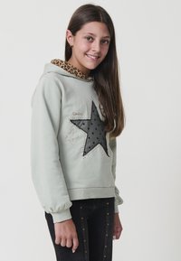 Chica sonriente con cabello largo y castaño, vistiendo una sudadera verde claro con un gran diseño de estrella negra y pantalones negros con tachuelas doradas.