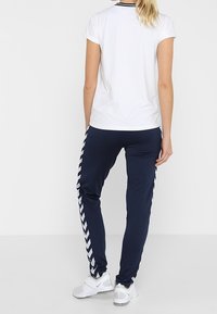 Weißes Kurzarmoberteil mit schwarzem Kragen, kombiniert mit marineblauen Jogginghosen mit einem weißen Chevronstreifen an der Seite, getragen mit weißen Turnschuhen.