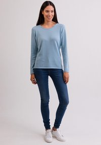 Ljusblå långärmad tröja, mjuk textur, rund halsringning, kombinerad med mörka slim fit jeans och vita sneakers, stående mot en neutral bakgrund.