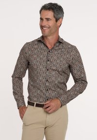 Mann lächelt und trägt ein gemustertes langärmliges Hemd mit geometrischen Formen und beigen Hosen, während er einen dunkelbraunen Gürtel anpasst.