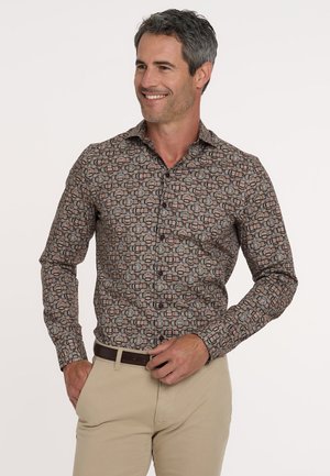 Man die glimlacht terwijl hij een gestreept lang-shirt met geometrische vormen en beige broeken draagt, hand die een donkerbruine riem afstelt.