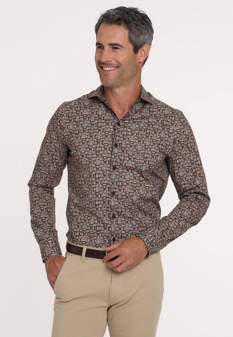 Mann lächelt und trägt ein gemustertes langärmliges Hemd mit geometrischen Formen und beigen Hosen, während er einen dunkelbraunen Gürtel anpasst.