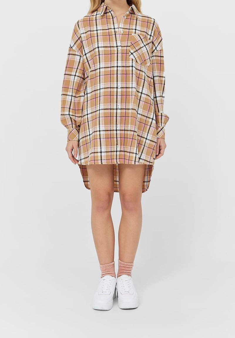 Femme portant une robe-chemise à carreaux oversize beige, blanche, rose et noire, manches longues, chaussettes roses et baskets blanches sur fond uni.