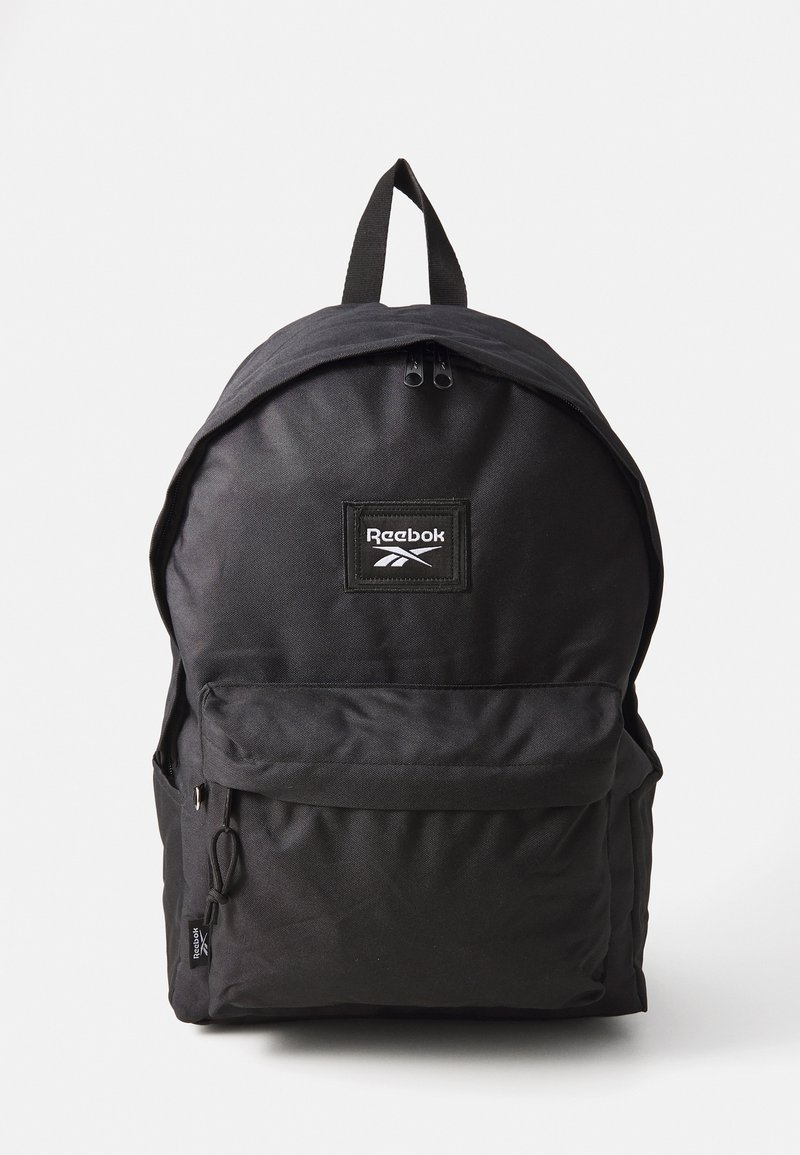 Schwarzer Stoffrucksack mit runder Form, ausgestattet mit einer Fronttasche, doppeltem Reißverschluss und einem oberen Griff. Reebok-Logo auf der Vorderseite.