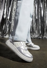 Sneaker argento e bianchi con una texture liscia, dotati di chiusura con lacci, strisce laterali e una suola bianca spessa. Indossati con jeans azzurri chiari.