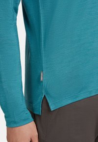 Teal long-sleeve shirt, lightweight fabric, rounded hem with side slits, reinforced stitching, and a small brand label on the side.