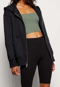 Svart zip-up hoodie i mjukt tyg, med en frontficka och ribbade ärmslut, lager på en grön crop top och svarta leggings.