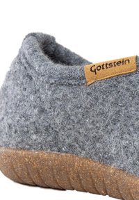Grau gefühlte Hausschuhe mit strukturiertem Obermaterial, runder Form und einer hellbraunen Sohle aus Naturkautschuk. Mit einem kleinen Lederetikett mit der Aufschrift "Gottstein."