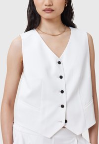 Weißes ärmelloses Tanktop mit V-Ausschnitt, ausgestattet mit sechs schwarzen Knöpfen, zwei Fronttaschen und einem glatten Stoff für eine maßgeschneiderte Passform.
