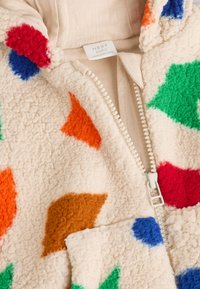 Veste bébé en polaire crème avec des formes pelucheuses colorées en orange, rouge, bleu, vert et marron. Dotée d'une fermeture éclair à l'avant et d'un détail de poche.
