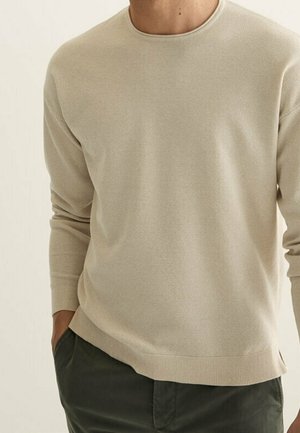 Homme portant un pull en maille beige à manches longues et un pantalon vert foncé, une main dans la poche, sur un fond clair uni.