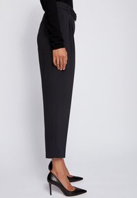 Musta muotoiltu housut, joissa on pleatit, lyhyt pituus, sileä pinta ja tiukka vyötärö, yhdistettynä teräväkärkisiin mustiin korkokenkiin.