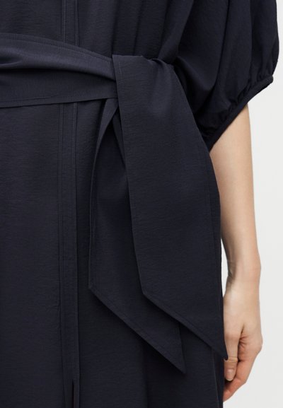 Κοντινό πλάνο ενός ατόμου που φοράει σκούρο μπλε φόρεμα με φουσκωτά κοντά μανίκια και μια φαρδιά υφασμάτινη ζώνη δεμένη σε φιόγκο στη μέση.