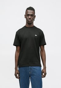 Juoda medvilninė marškinėliai su trumpomis rankovėmis, apvaliu iškirpimu ir mažu žaliu logotipu kairėje krūtinės pusėje. Dėvimi su mėlynomis džinsinėmis kelnėmis.