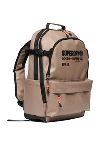 Beige ryggsäck med svarta remmar och accenter. Har logotyp, två fickor med dragkedja och dragkedjeskydd i orange. Slätt, hållbart material.