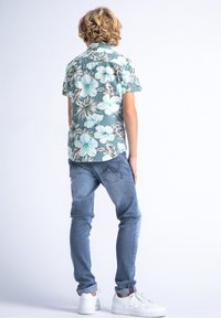 Camicia a maniche corte teal con motivo floreale, caratterizzata da fiori bianchi e verdi, abbinata a jeans azzurri chiari e sneakers bianche. Look casual e vestibilità rilassata.