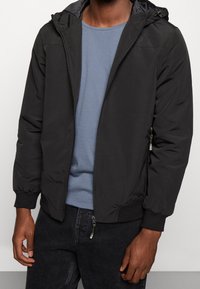 Mężczyzna w czarnej kurtce z kapturem na zamek, pod którą ma niebieską koszulkę z okrągłym dekoltem oraz czarne dżinsy, stoi na tle jednolitego tła.