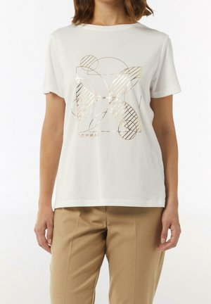 Femme portant un T-shirt blanc à manches courtes avec un motif géométrique doré de papillon, associé à un pantalon beige sur fond uni.