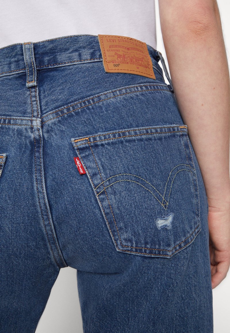 Jeans de mezclilla azul oscuro con un parche de cuero en la parte posterior, costuras en forma de arco en los bolsillos y una pequeña etiqueta roja visible cerca del bolsillo derecho.