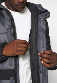 Szara ocieplana kurtka z zamkiem z przodu, wyposażona w wysoki kołnierz i elementy na rzepy. Wewnętrzna warstwa ma fakturę, pod spodem znajduje się prążkowany biały sweter.