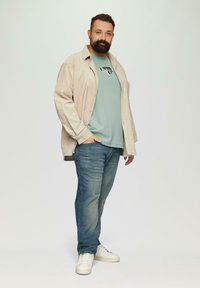 Beige corduroy overhemd over een lichtblauw T-shirt, blauwe spijkerbroek en witte sneakers; losse pasvorm en gelaagd ontwerp met een casual stijl.