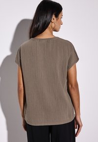 Khaki kurzärmliges Oberteil aus strukturierter, zerknitterter Stoff. Abgerundeter Saum und lockere Passform, kombiniert mit schwarzen Hosen. Keine sichtbaren Muster oder Akzente.