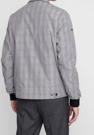 Mann trägt eine grau-weiß karierte Jacke mit schwarzen Bündchen und dunkelgraue Hose, von hinten vor einfachem Hintergrund gezeigt.