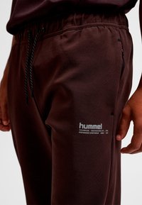 Brune sportsbukser med en tekstureret overflade, elastisk linning og justerbar snøre. Har hvidt logo på låret og sidelommer.