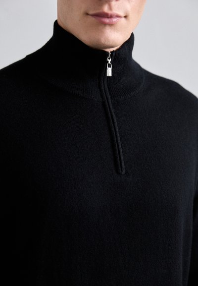 Schwarzer Pullover mit Reißverschluss aus weichem Stoff, mit hohem Kragen, einem halben Reißverschluss und einer glatten Textur ohne sichtbare Muster.