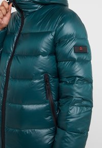 Giacca a vento trapuntata color verde acqua con tasche con zip nere e una piccola etichetta con logo sul braccio sinistro, indossata da una persona che aggiusta il colletto.
