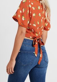 Hnědo-oranžový květinový crop top s krátkými rukávy, krajkovým okrajem a detailem s uvázáním na zádech, spárovaný s modrými džíny s vysokým pasem.