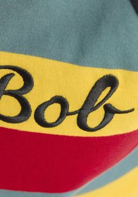 Približan posnetek tkanine z vodoravnimi turkiznimi, rumenimi in rdečimi črtami ter črnim vezenim pisanim besedilom »Bob«.