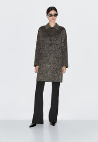Cappotto in stampa leopardo grigio e nero, con bottoni, colletto a camicia e maniche lunghe, indossato con pantaloni neri e occhiali da sole.