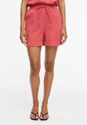 Kvinne iført korallrosa teksturerte shorts med knyttet midje og svarte thong-sandaler, stående mot hvit bakgrunn.