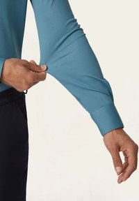 Lange mouw shirt in teal, gemaakt van soepel fabric. De manchetten zijn met een knoop gesloten en hebben een getailleerde pasvorm. Een hand grijpt de elastische textuur van de mouw.